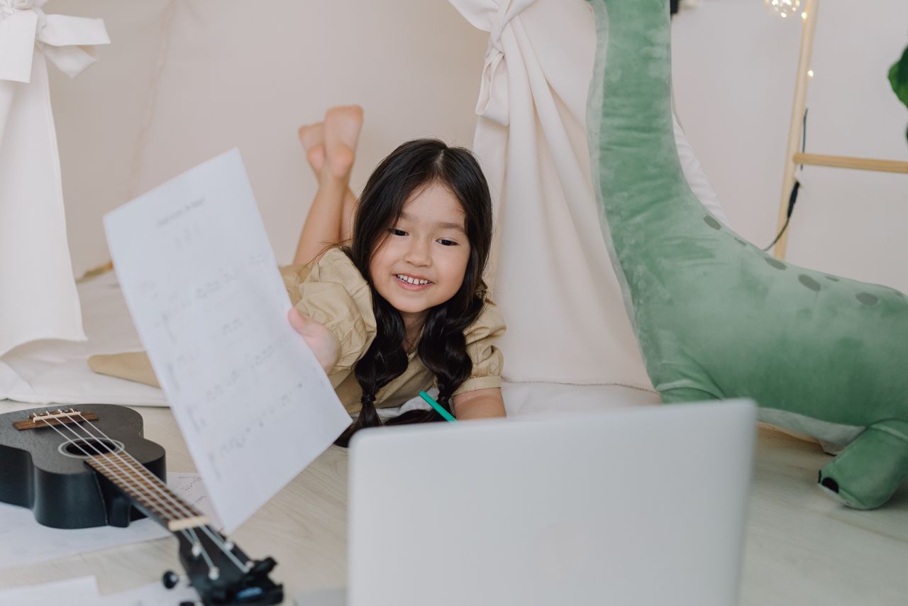 Happy child learning ukulele online at home with sheets and laptop, showing engaging childhood education.